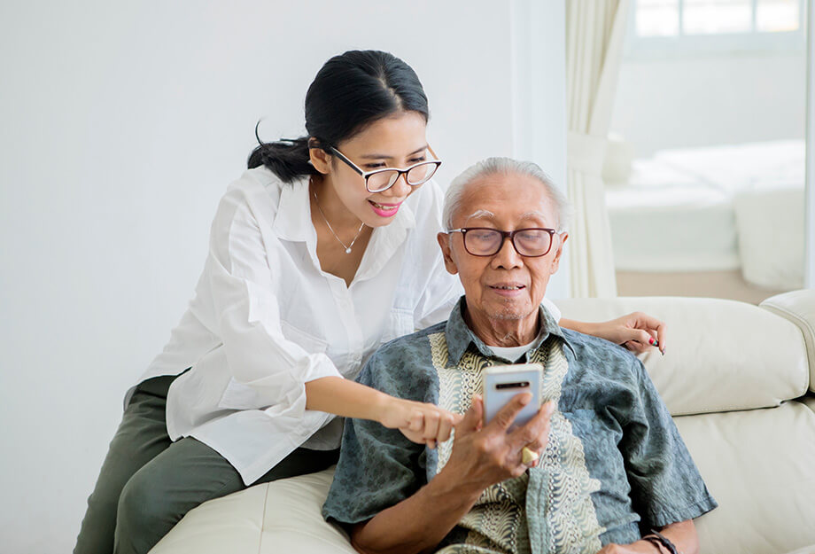 Young woman helping elderly man with smartphone in a living room setting.