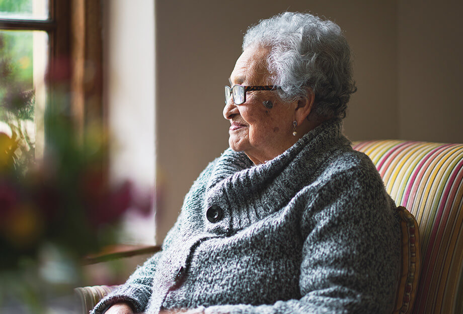 Elderly woman with glasses sitting by a window in a cozy unit, looking outside.