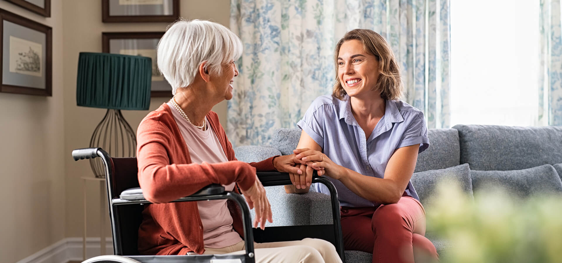 Senior woman in wheelchair chatting with a younger woman on the couch in a unit.