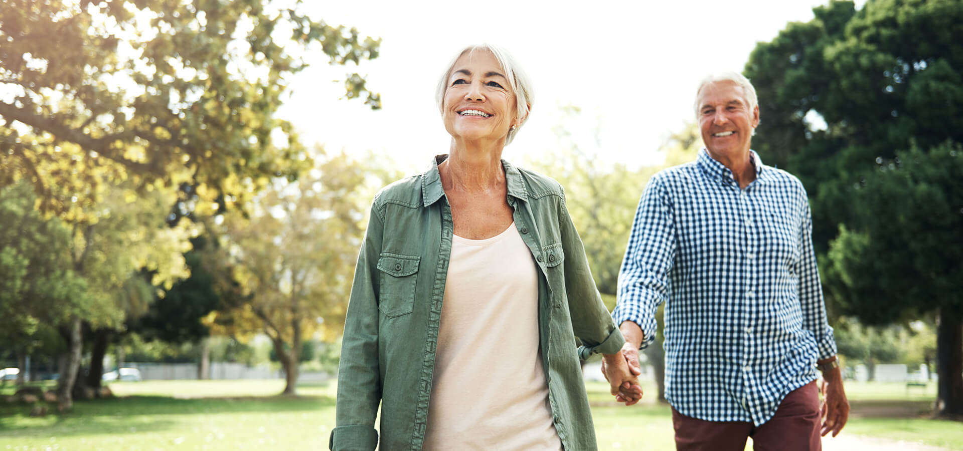 Elderly couple walking hand in hand in a sunlit park, smiling joyfully.