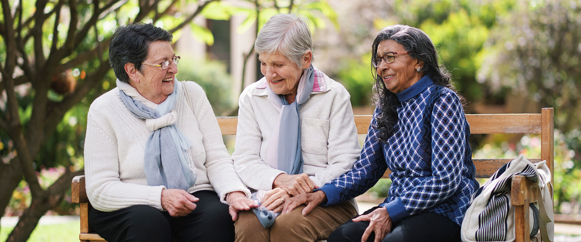 Three senior women laughing together on a bench in a garden setting.