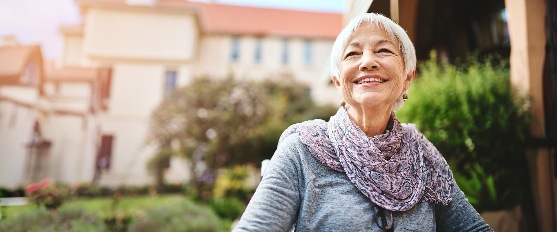 Elderly woman smiling outside, with a building and greenery in the background.