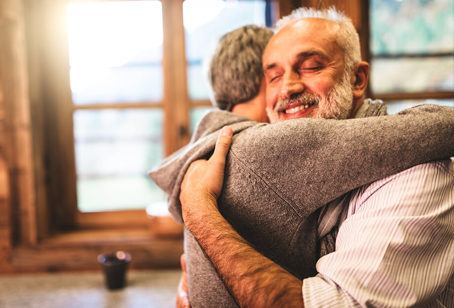 Two older adults embracing warmly in a cozy room with large windows.